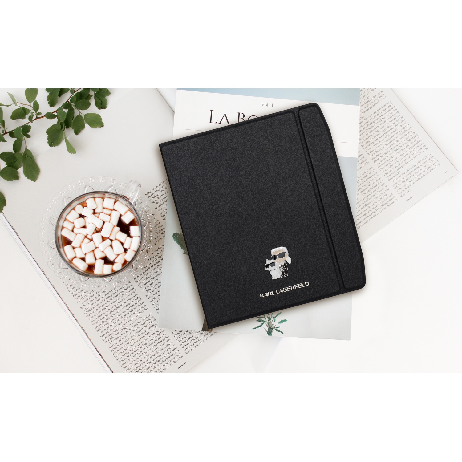 Zwarte Karl Lagerfeld tablet hoes, een kop marshmallow warme chocolademelk, tijdschriften en groene takjes op witte tafel.