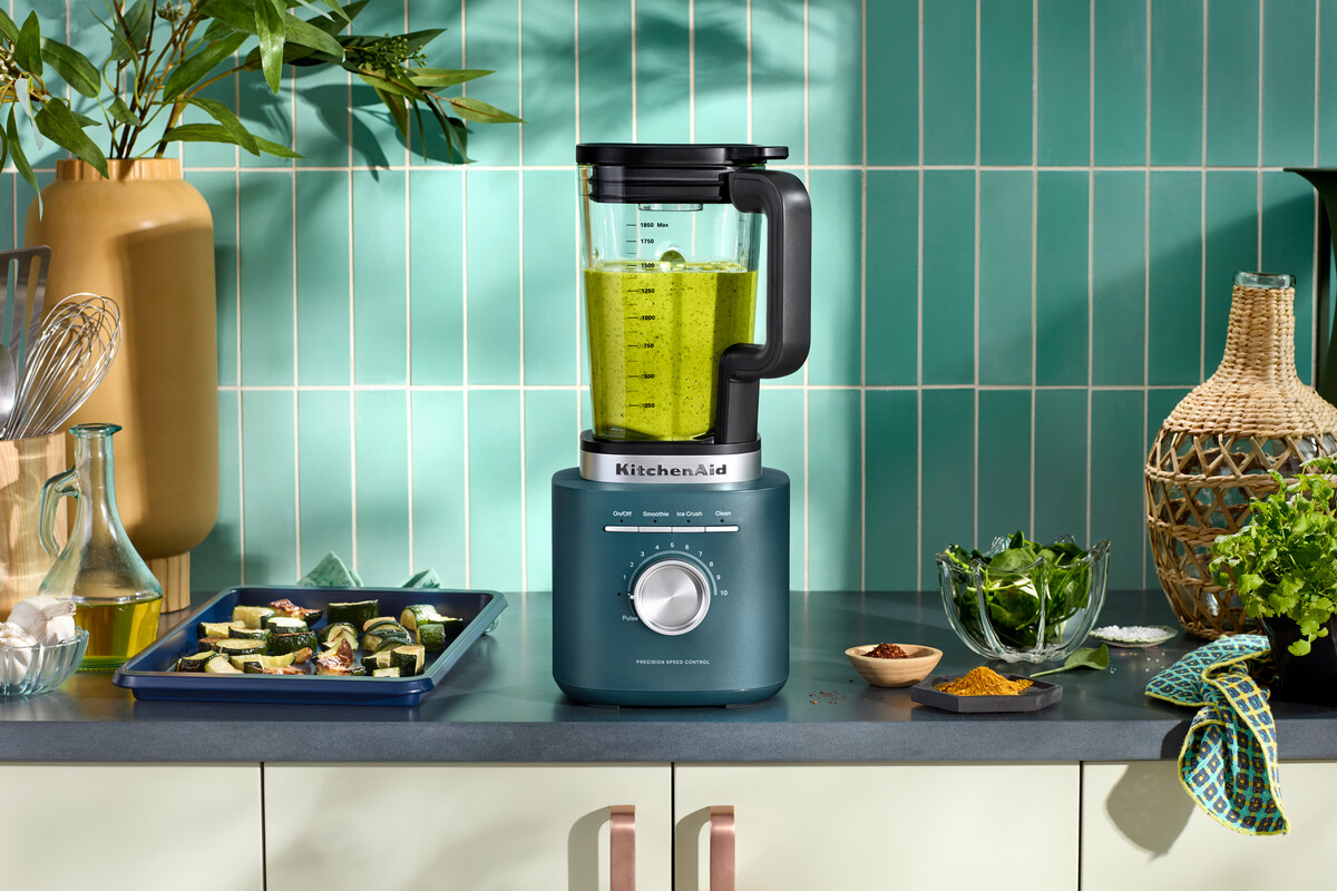 Ein blauer KitchenAid-Standmixer steht auf einer Küchentheke mit grünem Gemüse und Fliesenwand.