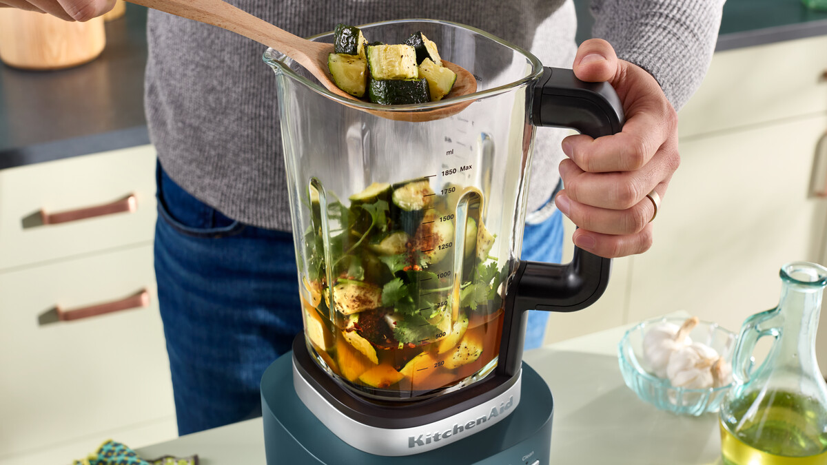 Person gibt mit einem Holzlöffel gehackte Zucchini in einen KitchenAid-Standmixer mit Zutaten.