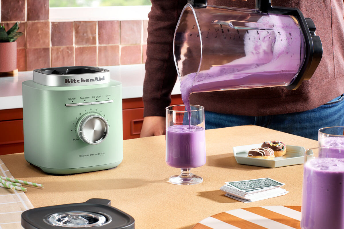 Eine Person gießt einen violetten Smoothie aus einer Mixerkanne in ein Glas auf einem Tisch.