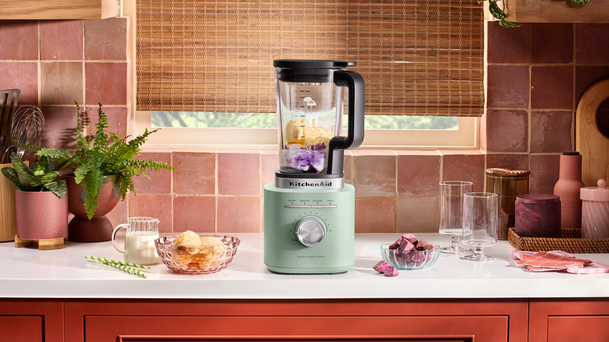 Ein pistaziengrüner KitchenAid-Blender steht auf einer Küchentheke mit Zutaten wie Eiscreme und violetten Früchten.