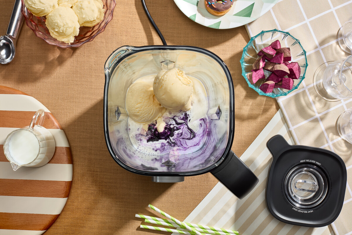 Ein Mixer mit Eiscreme und violettem Püree, umgeben von Zutaten wie Milch, Taro und Eiscreme auf einem Tisch.