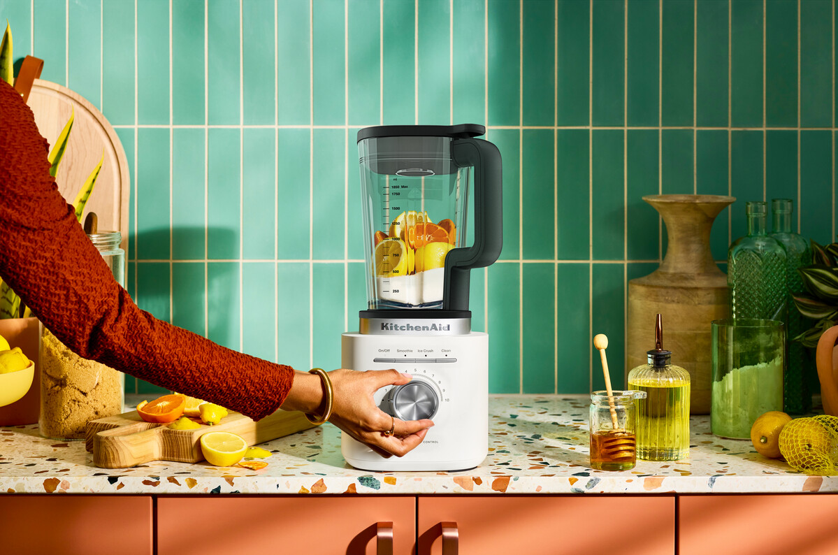 Eine Hand bedient einen weißen KitchenAid-Mixer gefüllt mit Zitrusfrüchten und Milch auf einer bunten Arbeitsplatte.