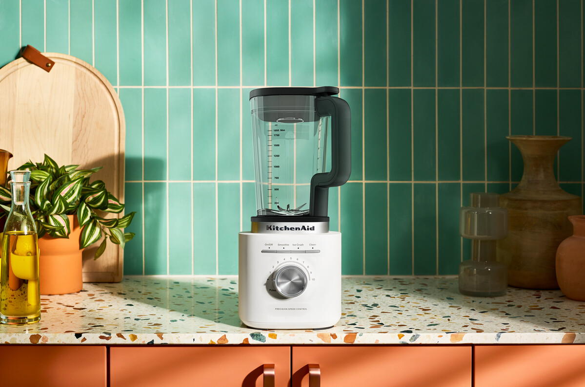 Ein weißer KitchenAid-Mixer steht auf einer Terrazzo-Arbeitsplatte vor grünen Fliesen mit Pflanzen und Vasen.