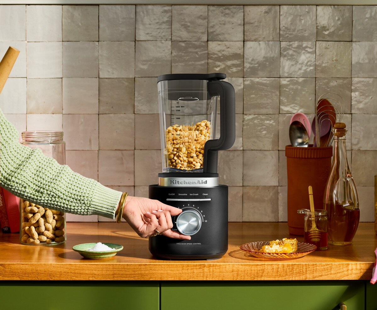 Eine Hand stellt einen schwarzen KitchenAid-Mixer ein, der mit Nöten gefüllt ist, auf einer Holzarbeitsplatte.
