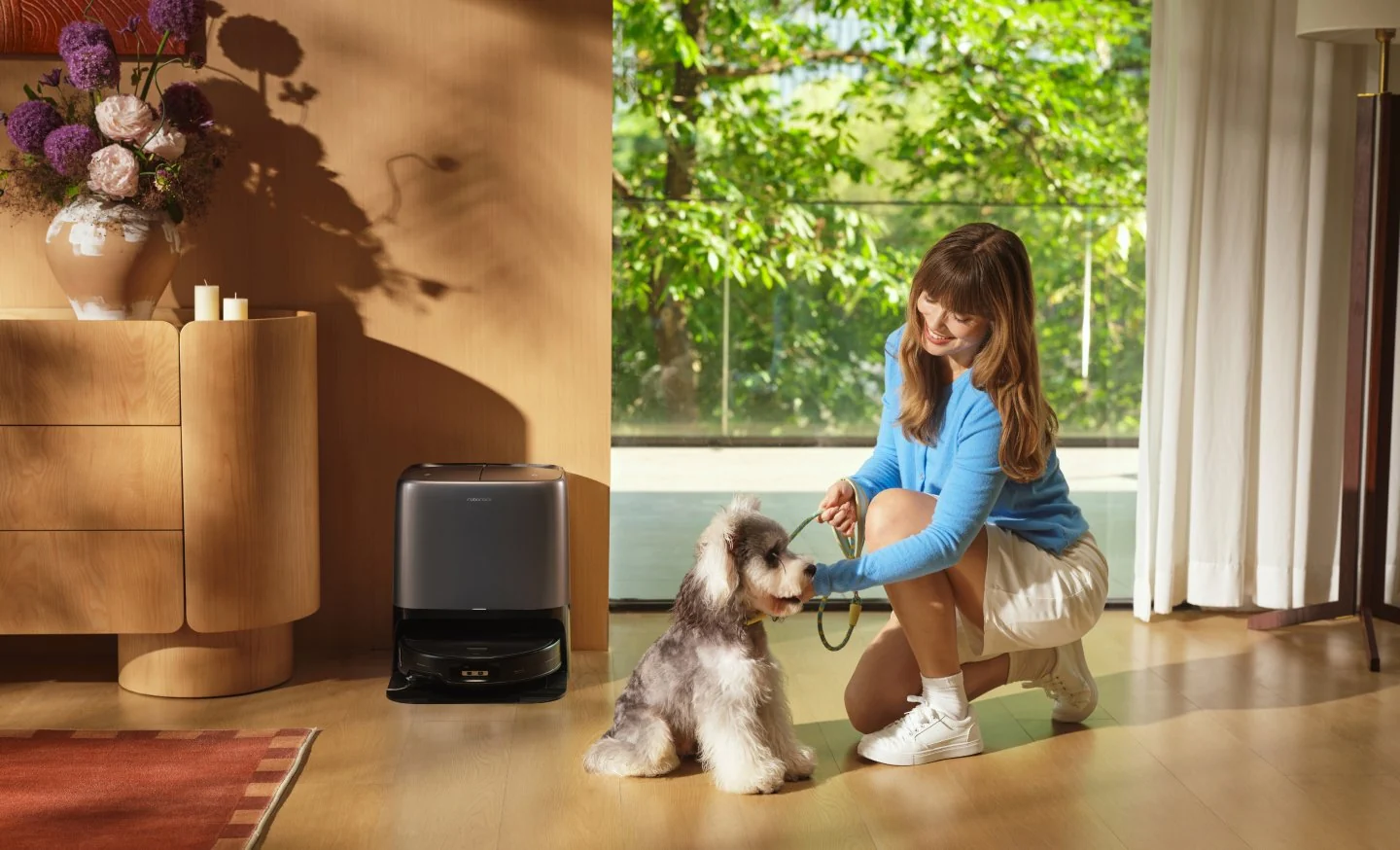 Une femme en haut bleu tient la laisse d'un petit chien à l'intérieur, avec un robot aspirateur à proximité.