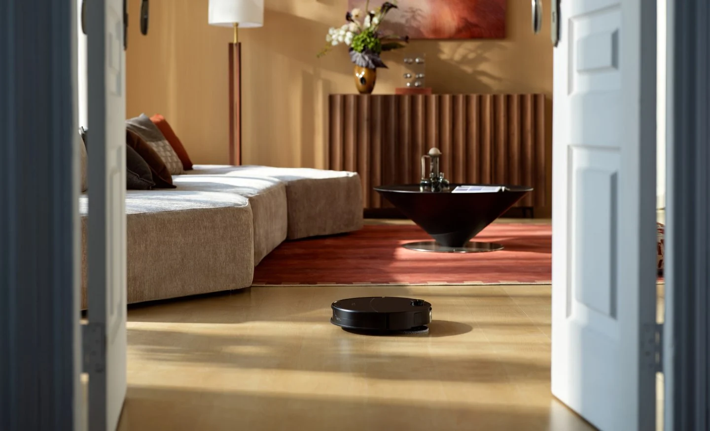 Un robot aspirateur noir nettoie un parquet dans un salon moderne et chaleureux vu à travers des portes blanches.