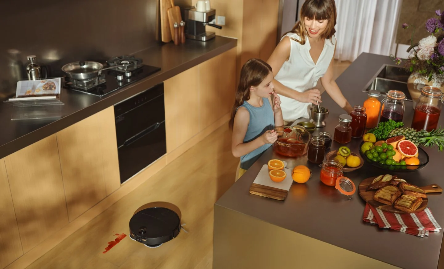 Une mère et sa fille préparent des plats dans une cuisine pendant qu'un robot aspirateur nettoie le sol.