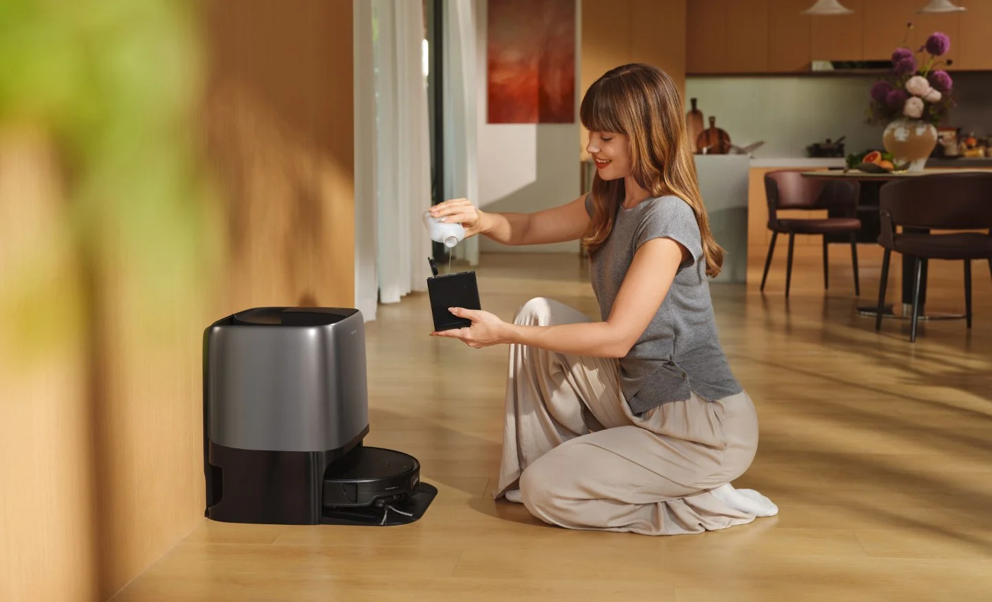 Femme agenouill ée sur un sol en bois verse du détergent dans le réservoir d'une station de robot aspirateur.