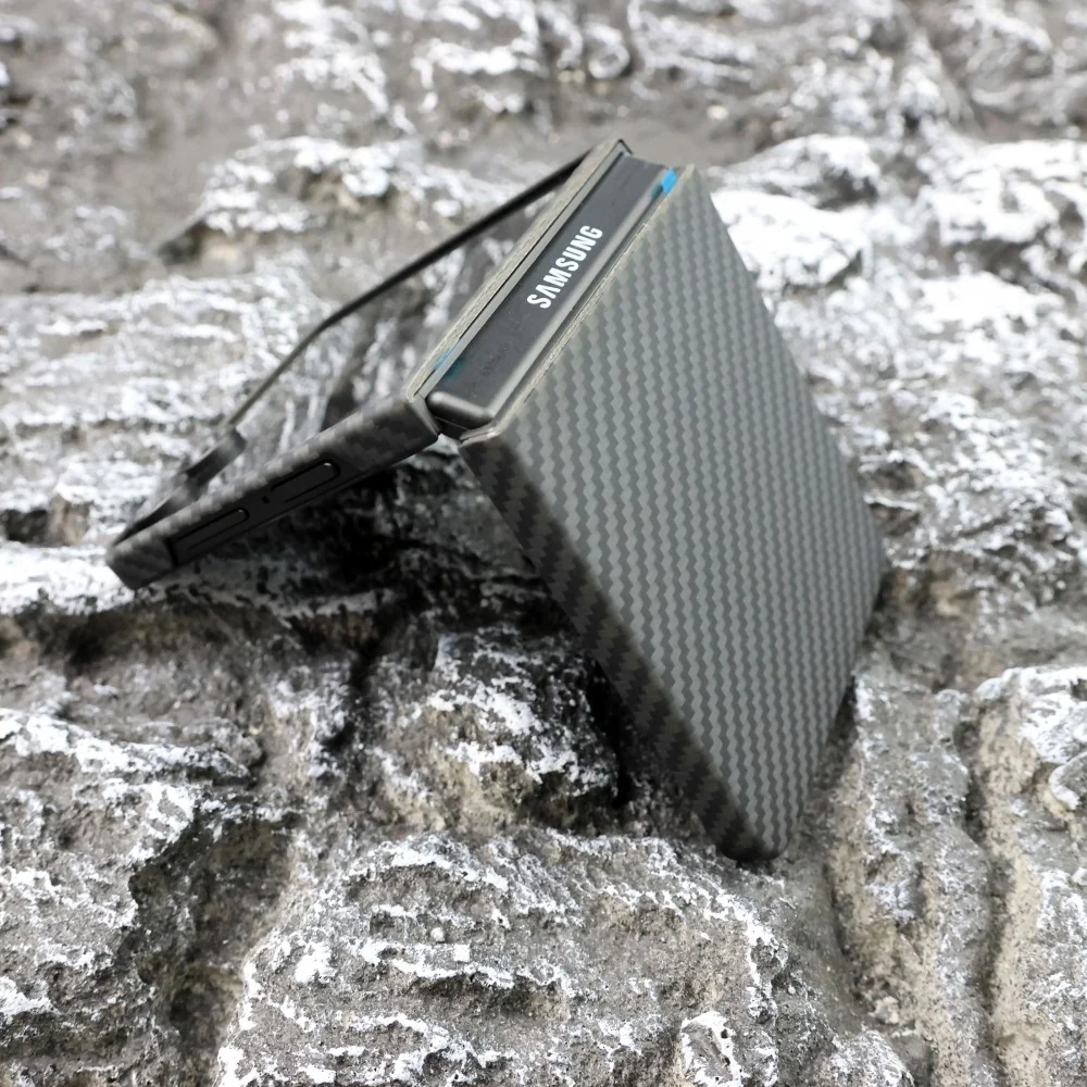 Félig nyitott, szénszálas mintázatú Samsung összecsukható telefon fekszik egy egyenetlen, szürke kőfelületen.