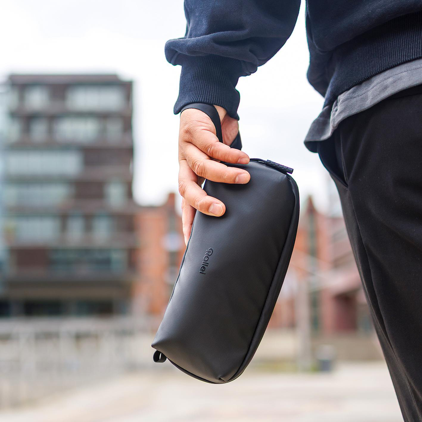 Eine Person hält eine kleine, schwarze Rollei-Tasche an einer Schlaufe, mit unscharfem städtischem Hintergrund.