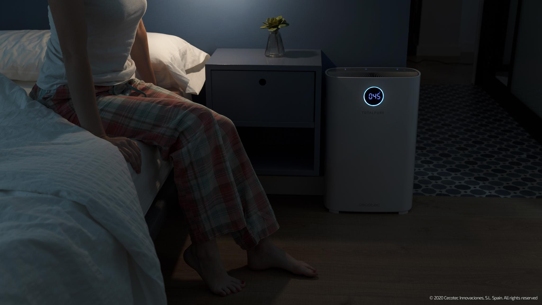 Una mujer se sienta en la cama en un dormitorio, con un purificador de aire blanco al lado.