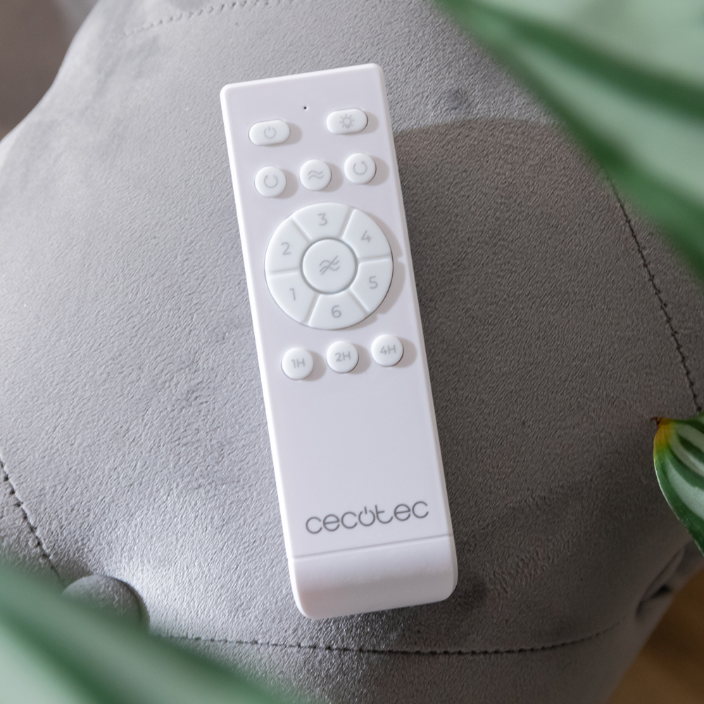 Un control remoto blanco de Cecotec con botones redondos descansa sobre una silla gris, enmarcado por hojas verdes.