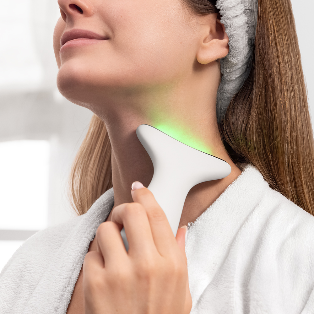 Una mujer en albornoz se masajea el cuello con un dispositivo blanco con luz verde brillante.