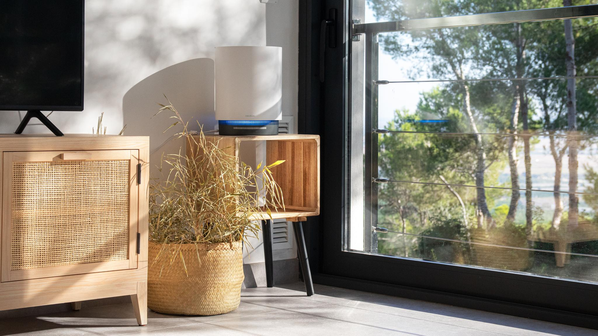 Un purificador de aire blanco con luz azul se encuentra en una mesa de madera junto a una ventana.