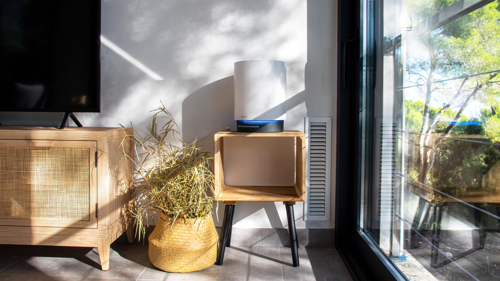 Un purificador de aire blanco con luz azul se encuentra en una mesa de madera al lado de una planta en maceta.