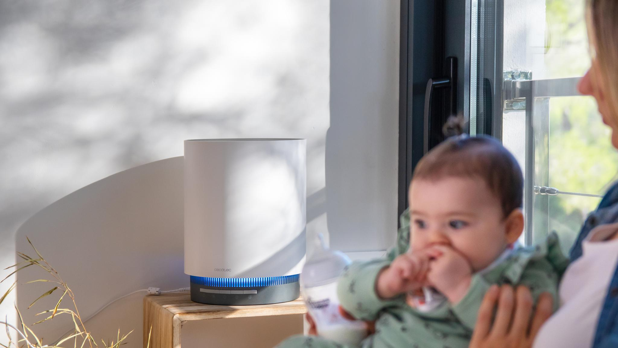 Un purificador de aire blanco con luz LED azul se encuentra en una mesa de madera junto a un niño y una ventana.