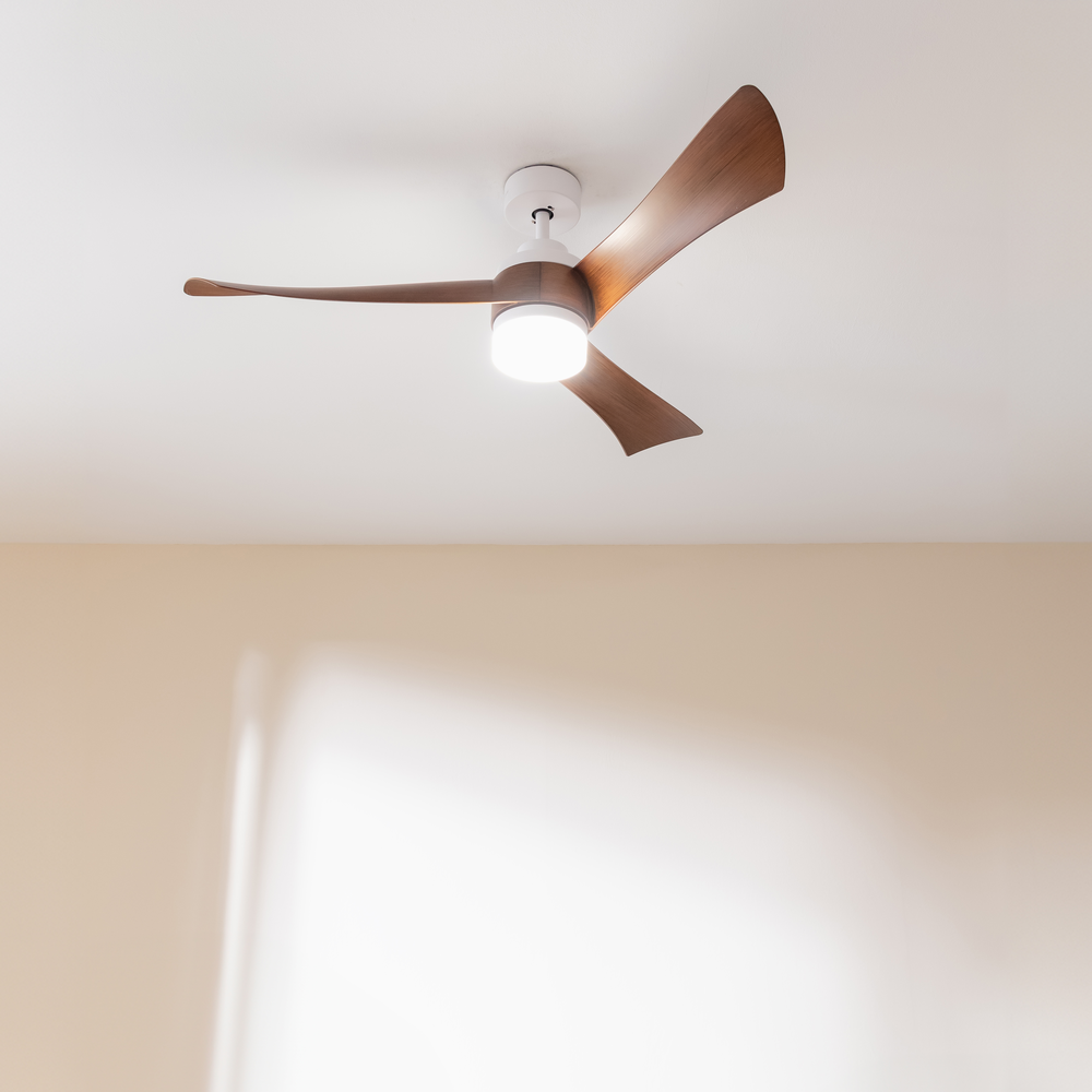 Un ventilador de techo moderno con tres aspas de madera y una lámpara iluminada montado en un techo blanco.
