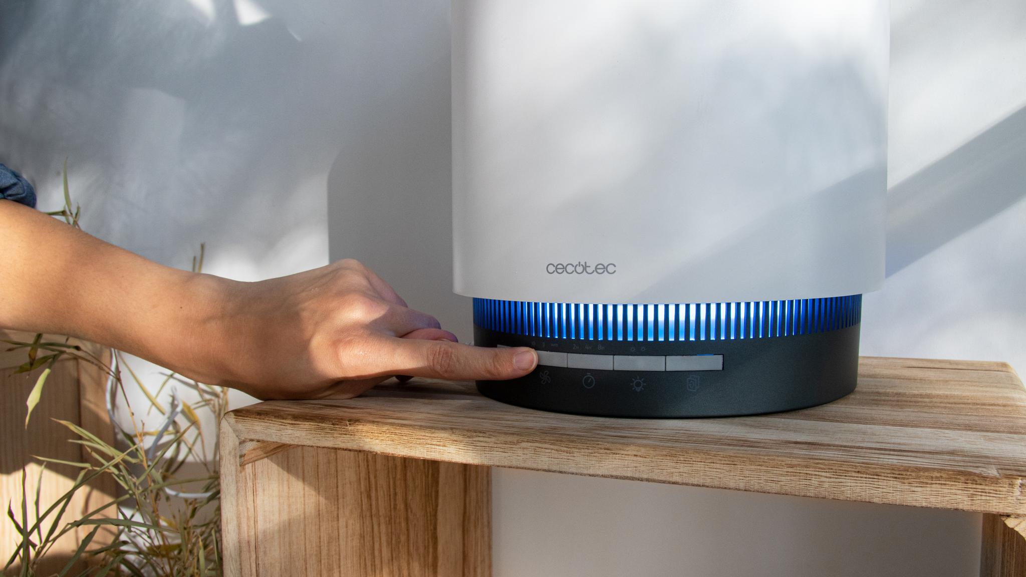 Una mano opera el dispositivo Cecotec blanco y negro en una balda de madera, con luz azul.