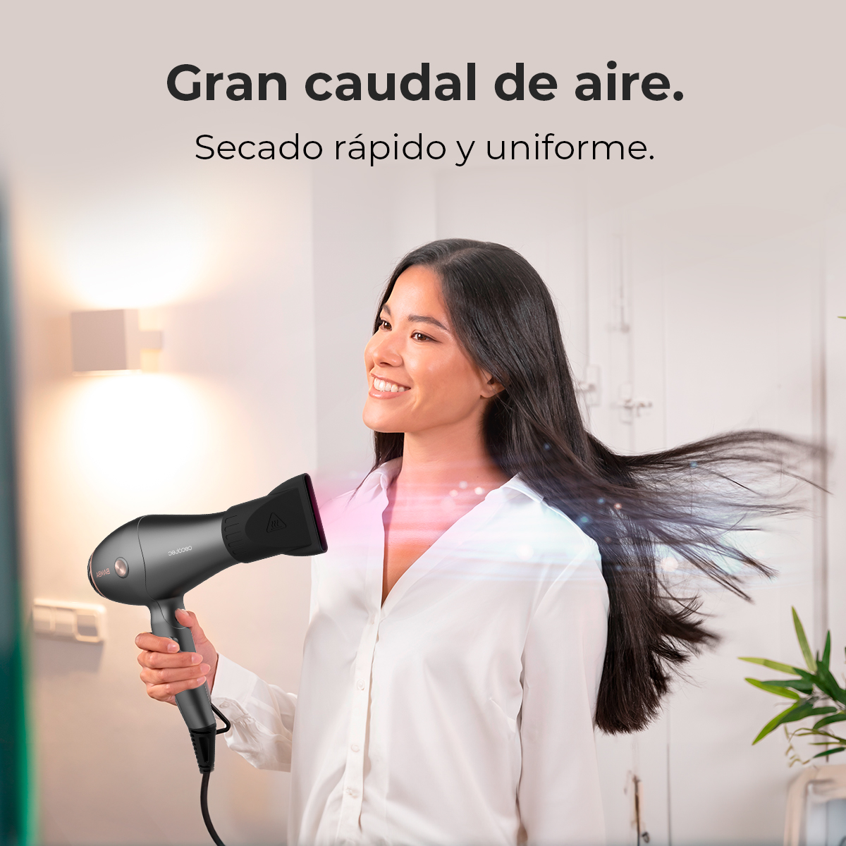 Una mujer sonriente seca su largo cabello negro con un secador gris moderno en una habitación luminosa.