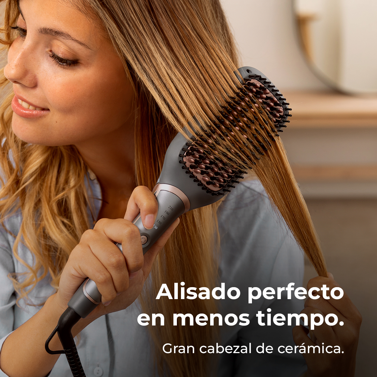 Una mujer alisa su cabello rubio con un cepillo alisador gris y oro rosa en una habitación luminosa.