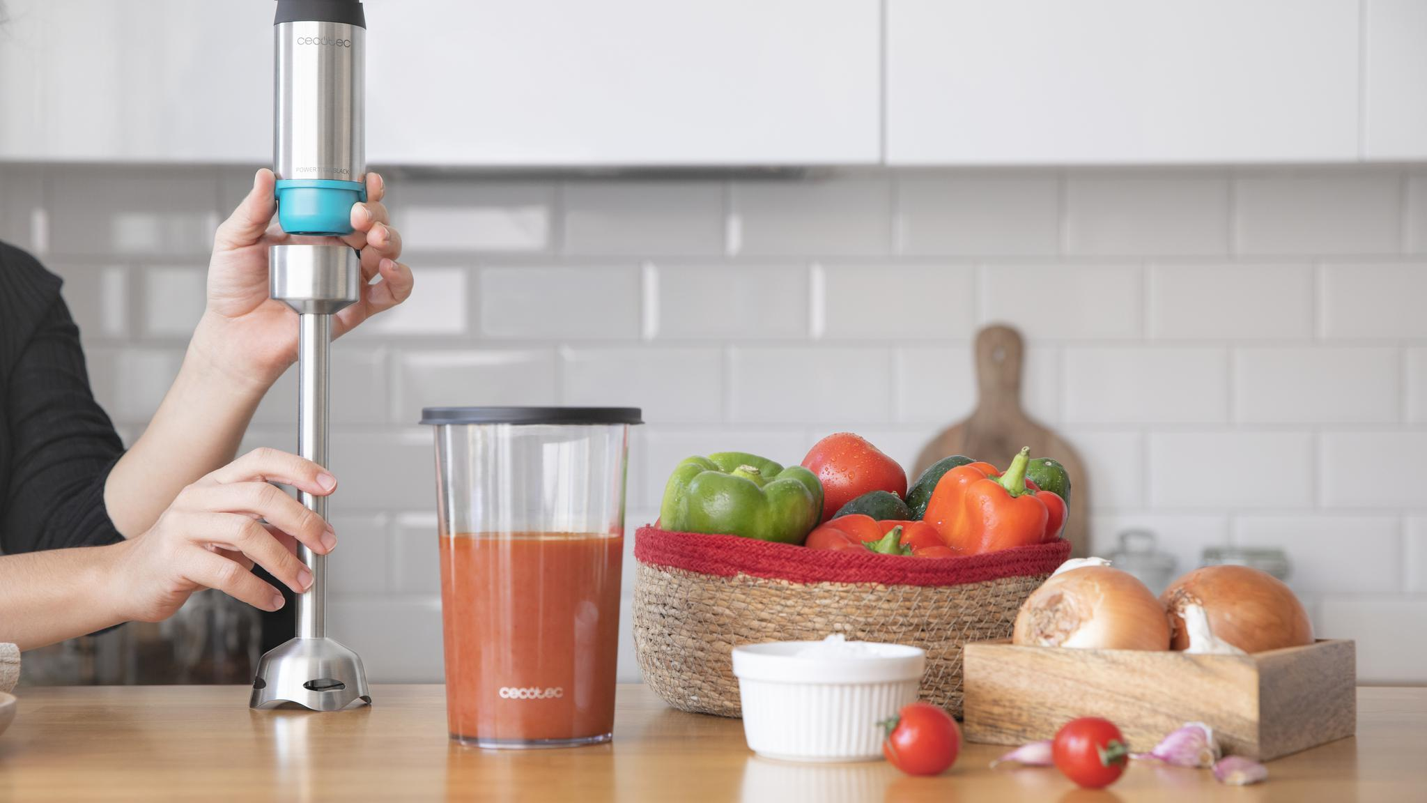 Una persona ensambla una batidora de mano en una encimera de cocina con verduras frescas y un recipiente con jugo.