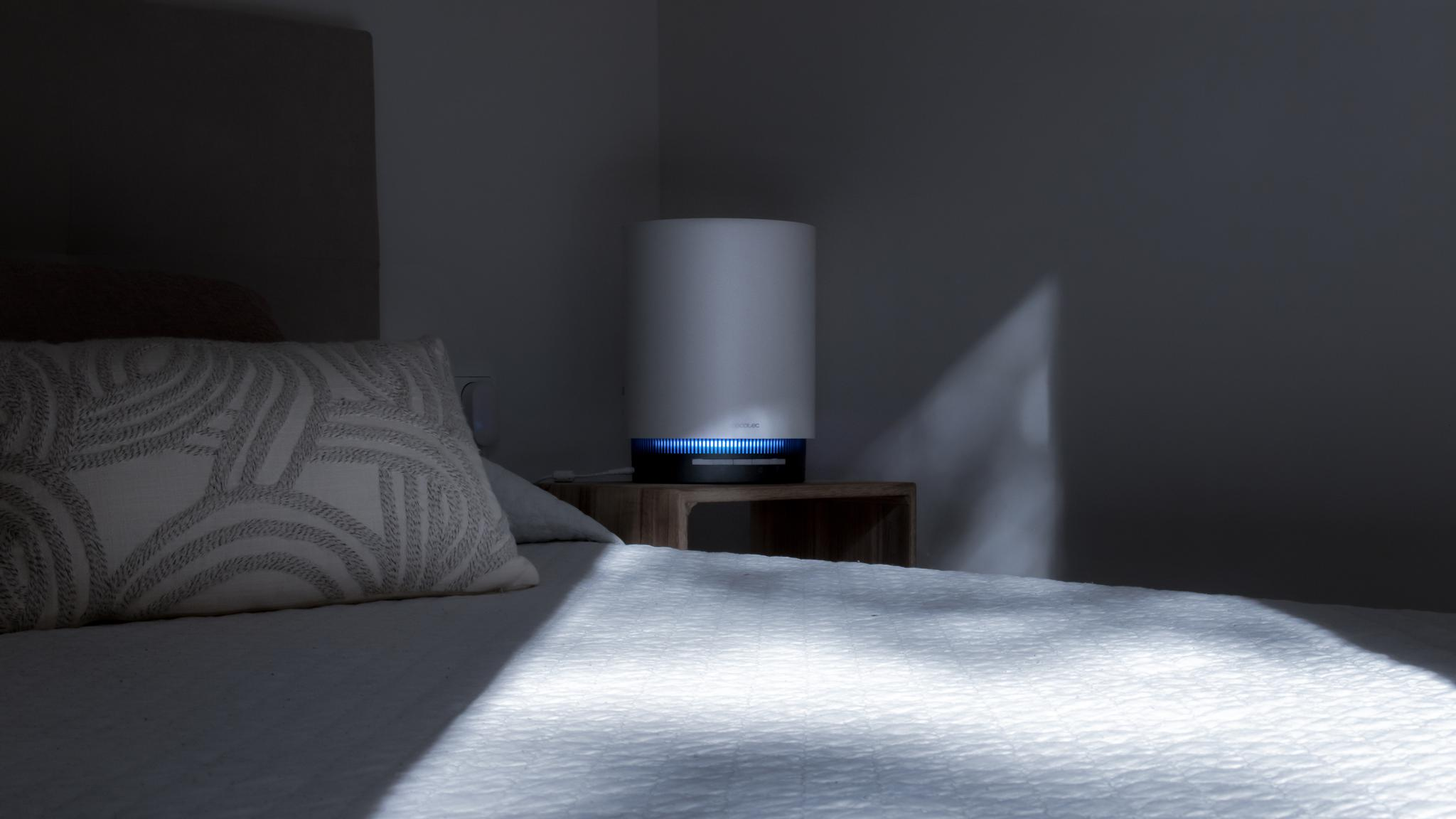 Un purificador de aire blanco con luz azul sobre una mesita de noche junto a la cama.