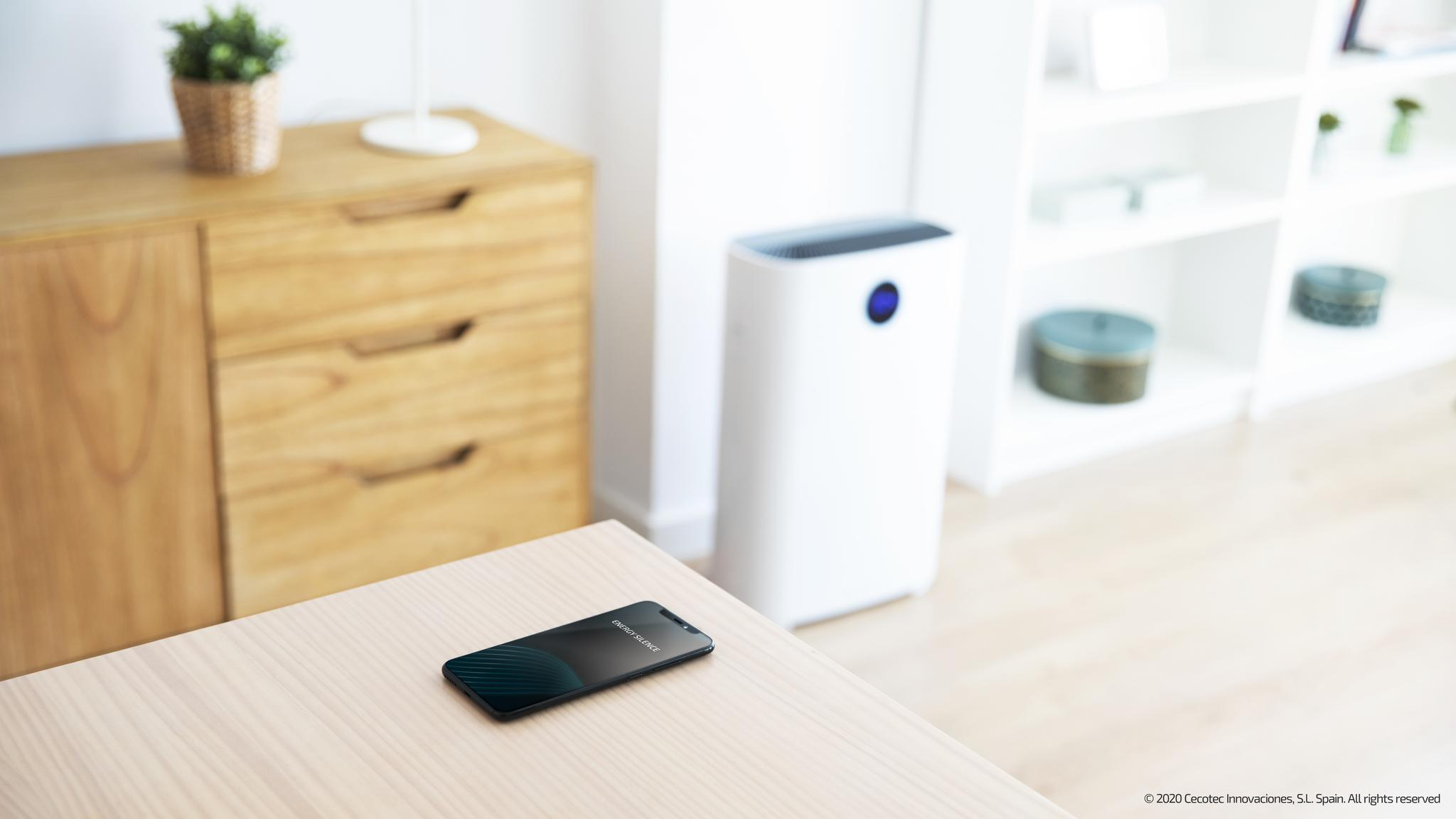 Un smartphone descansa sobre una mesa de madera en una habitación luminosa, con un purificador y una estantería.
