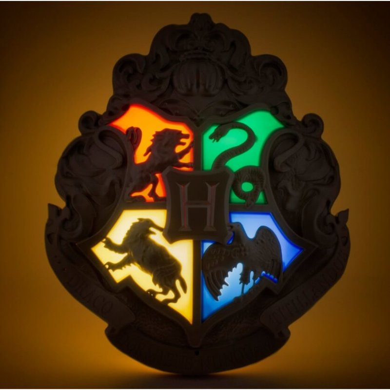 Un aplique de la escuela Hogwarts iluminado sobre fondo oscuro, con cuatro secciones de colores y una H central.