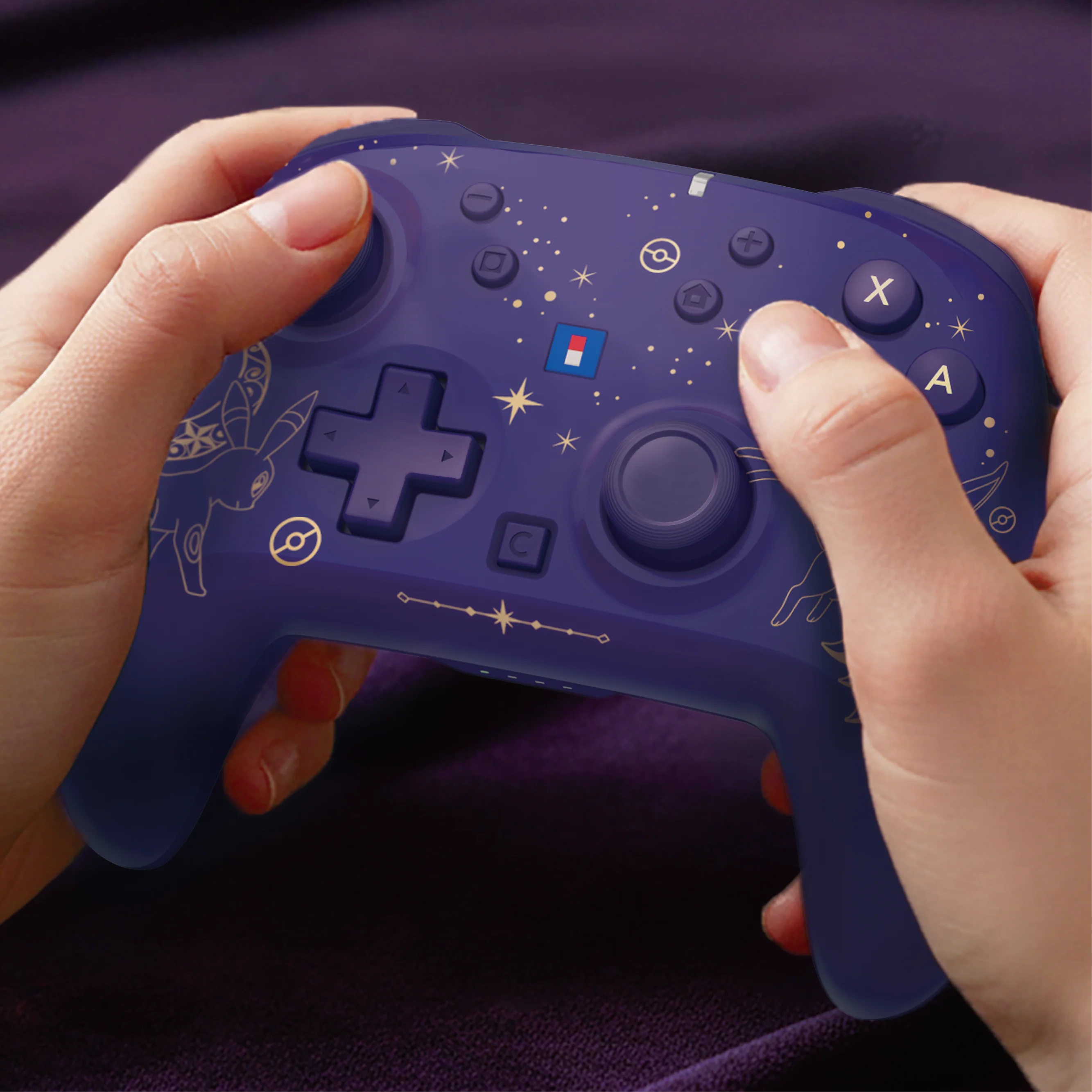 Mani che tengono un controller di gioco wireless blu con motivi di stelle e contorni di pokemon dorati su tessuto viola.