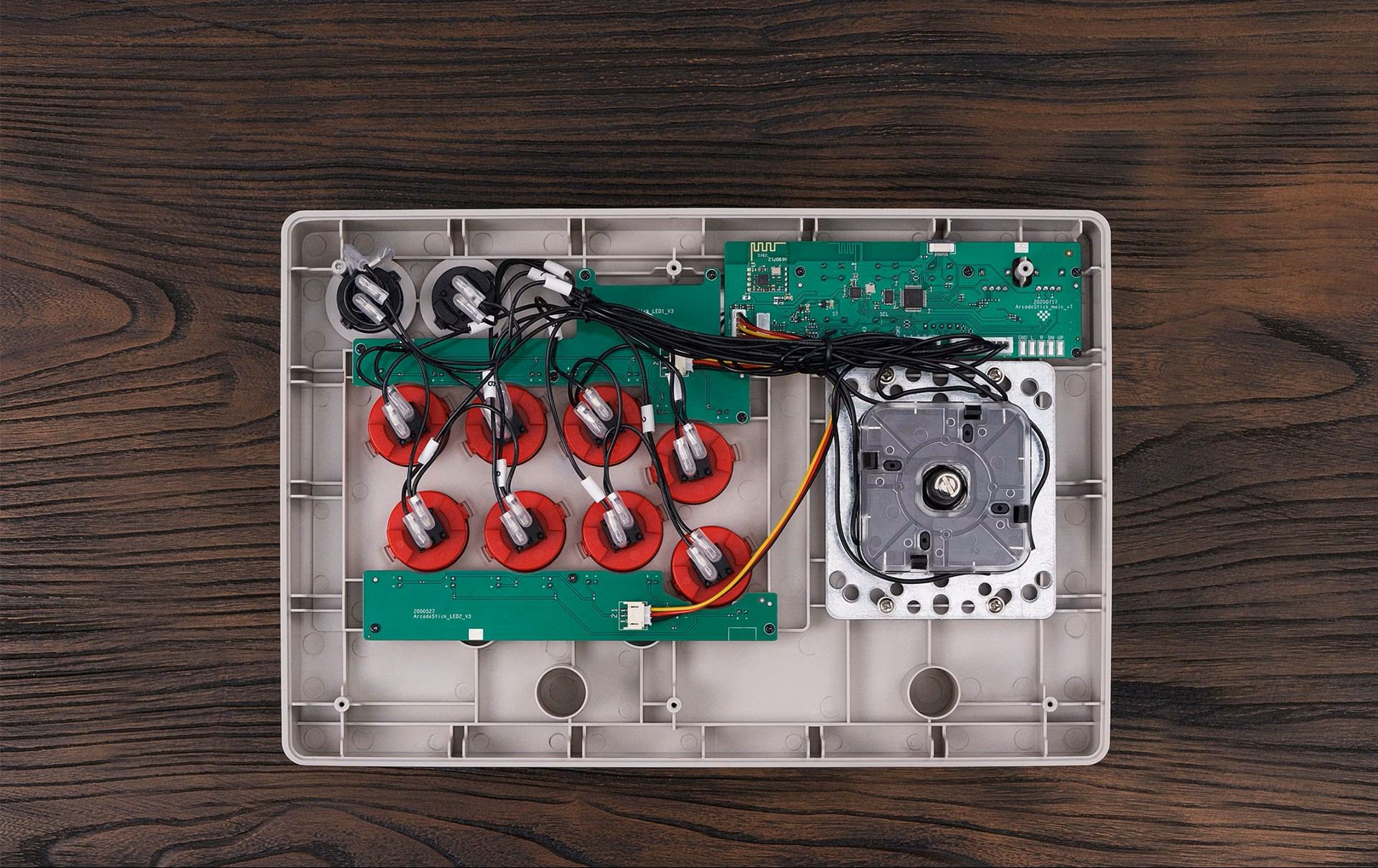 Interno di un joystick arcade con scheda verde, pulsanti rossi e cavi sul legno.