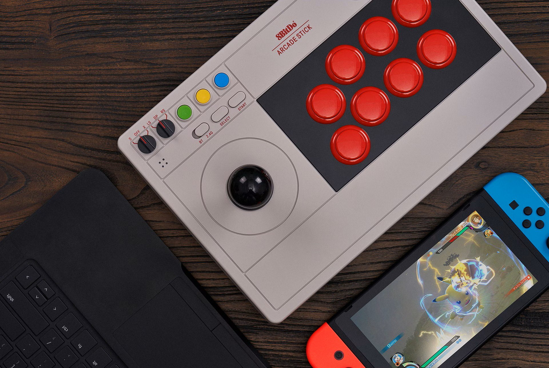 Joystick arcade grigio 8BitDo, una console Nintendo Switch e una tastiera nera su legno.