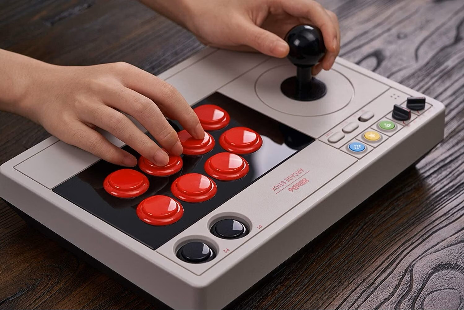 Mani che giocano su un joystick arcade grigio retro con pulsanti rossi su un tavolo di legno.