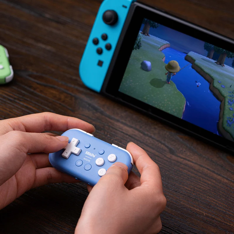 Un mini gamepad blu 8BitDo è tenuto in mano davanti a una console Nintendo Switch che mostra un videogioco.
