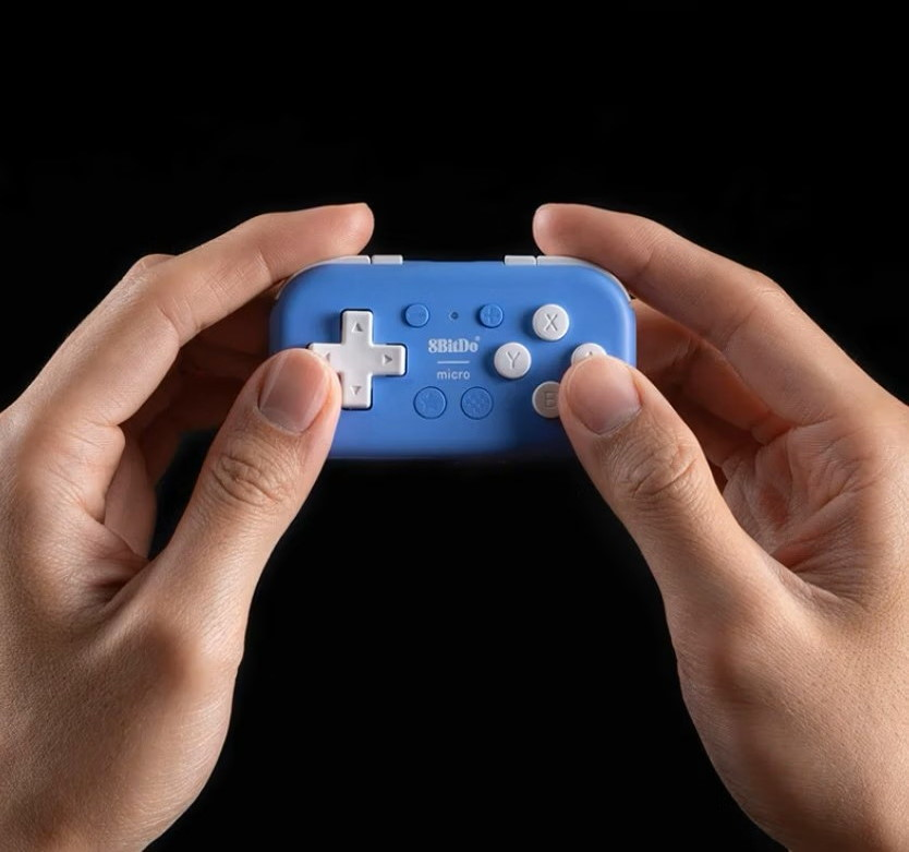 Una persona tiene in mano un piccolo controller di gioco blu 8BitDo micro con tasti bianchi su sfondo nero.