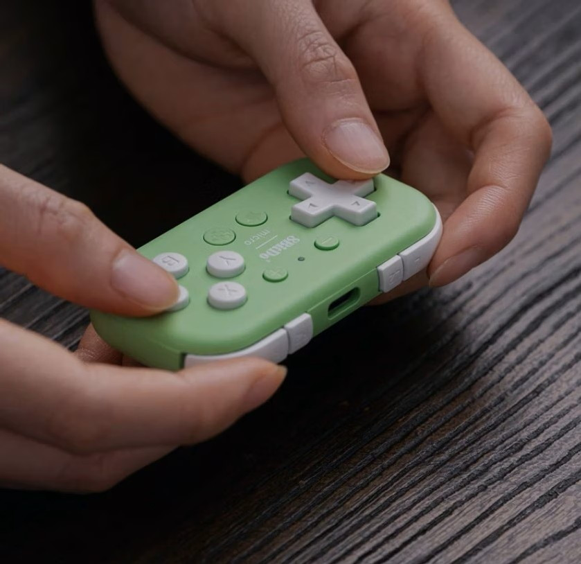 Mani che tengono un piccolo gamepad Bluetooth 8BitDo micro verde chiaro e bianco su un tavolo di legno scuro.