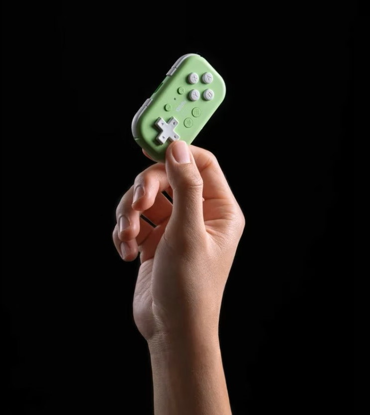 Una mano tiene un piccolo controller di gioco verde con pulsanti bianchi su sfondo nero.