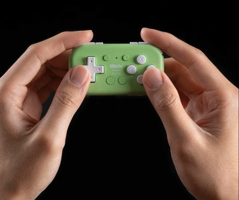 Due mani tengono un piccolo controller di gioco 8BitDo Micro verde chiaro su sfondo scuro.