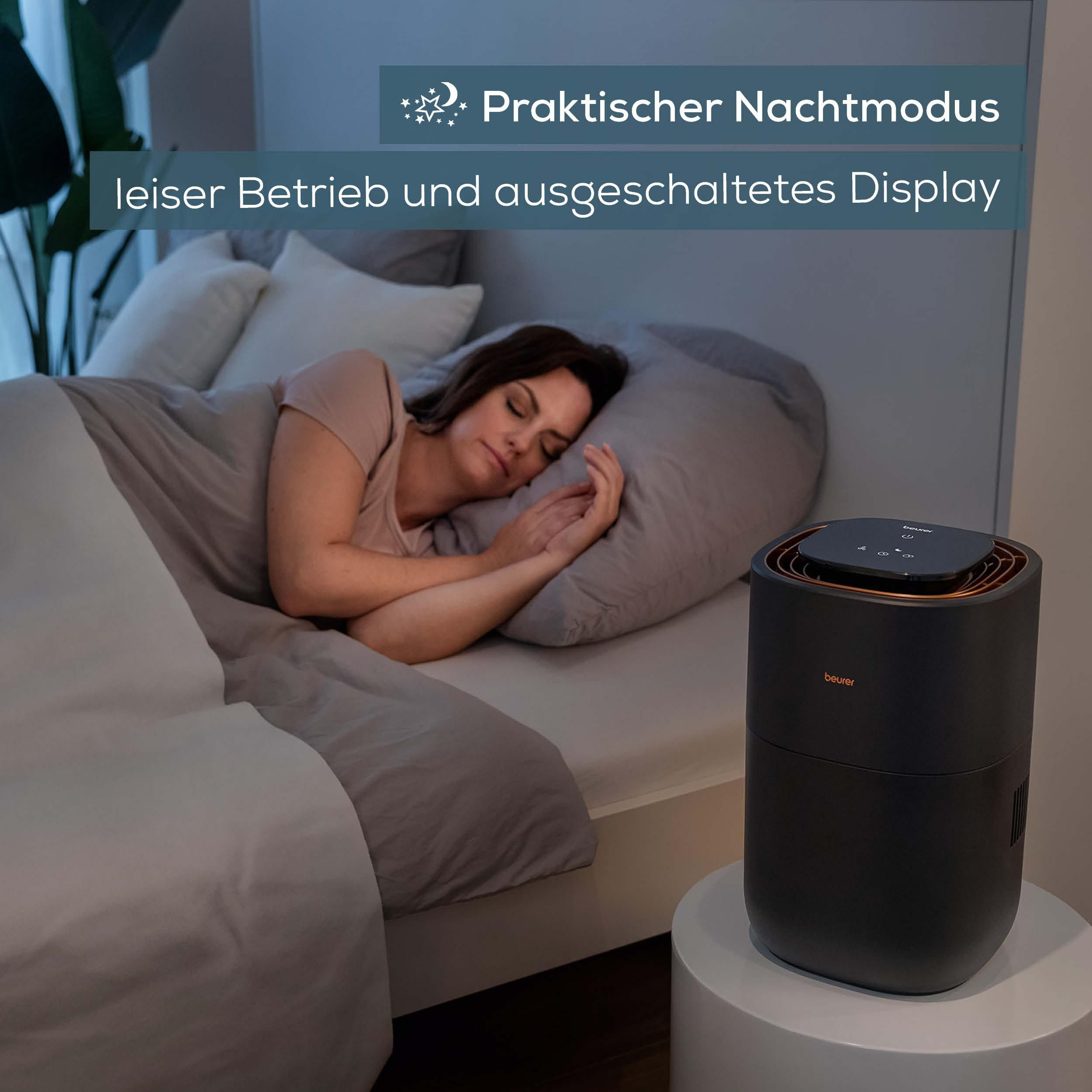 Eine Frau schläft friedlich im Bett, daneben steht ein schwarzes Beurer-Gerät auf einem runden Tisch.
