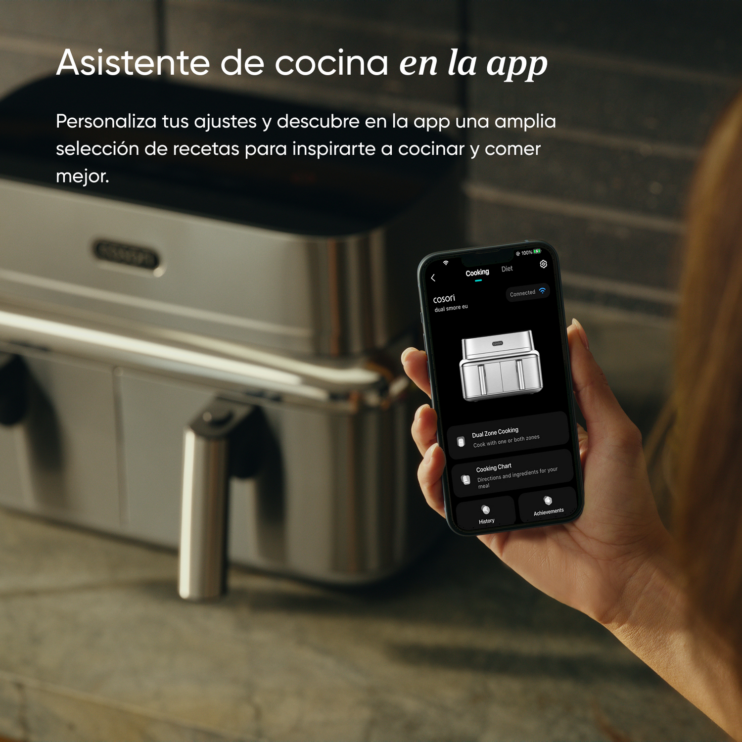 Una mano sostiene un smartphone con una aplicación de cocina para una freidora de aire plateada al fondo.