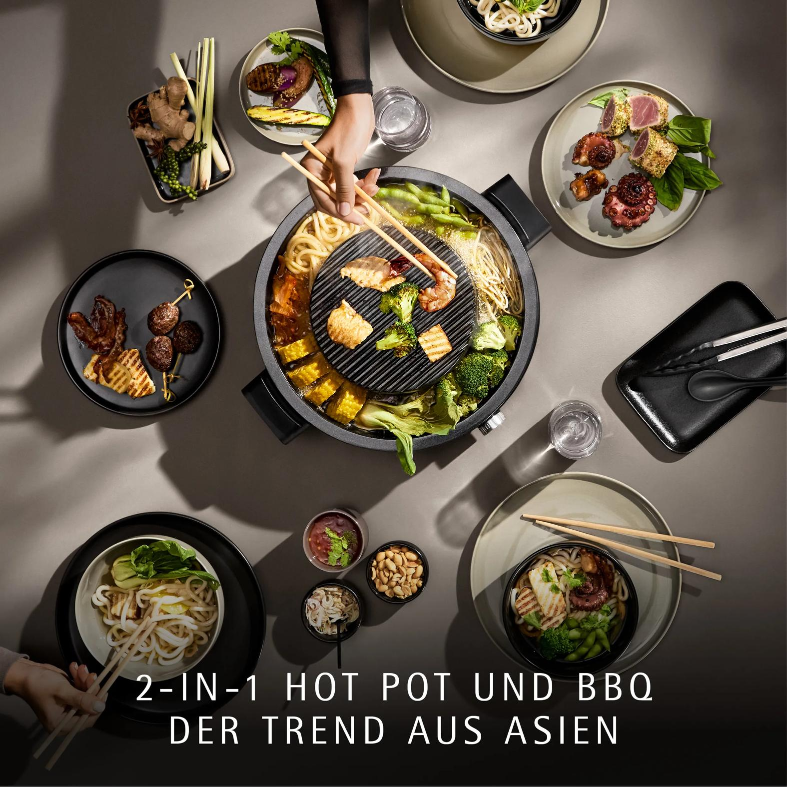 Top-Ansicht eines 2-in-1-Hot-Pot-und-BBQ-Grills mit verschiedenen Beilagen auf Tellern und Händen mit Stäbchen.