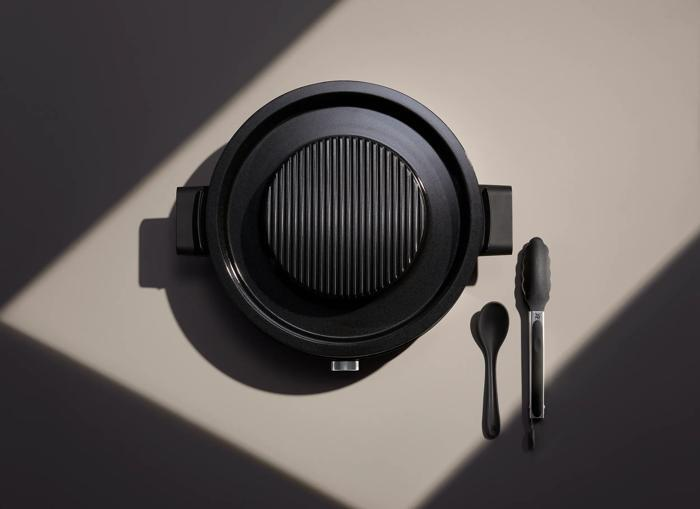 Eine Draufsicht auf einen schwarzen Elektrogrill mit Löffel und Zange auf einer zweifarbigen Oberfläche.