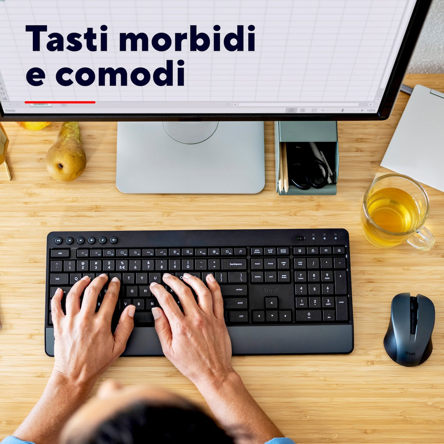 Vista dall'alto di mani che digitano su una tastiera nera, vicino a monitor, mouse e tazza di tè su una scrivania.