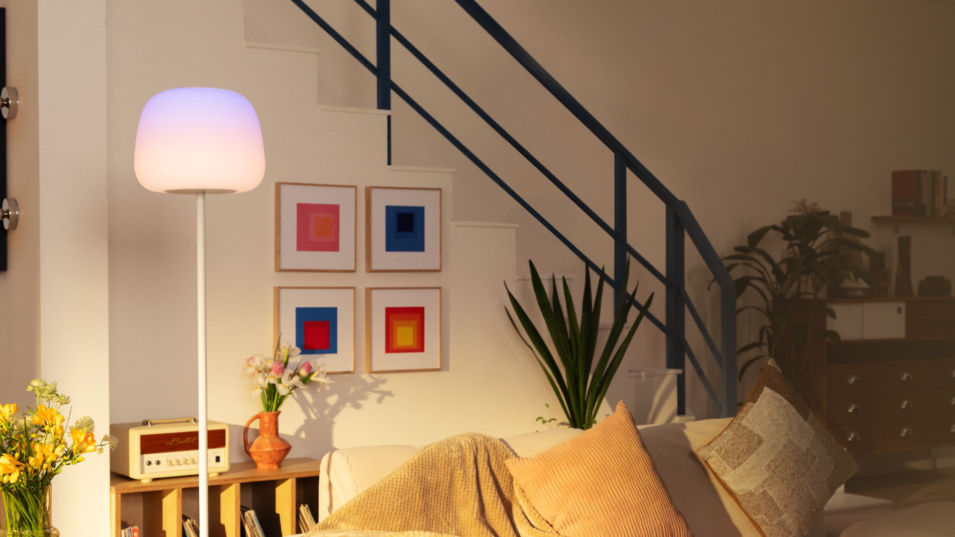 Wohnzimmer mit heller Stehlampe, Sofa, farbigen Bildern an der Wand und Treppe mit dunklem Geländer.