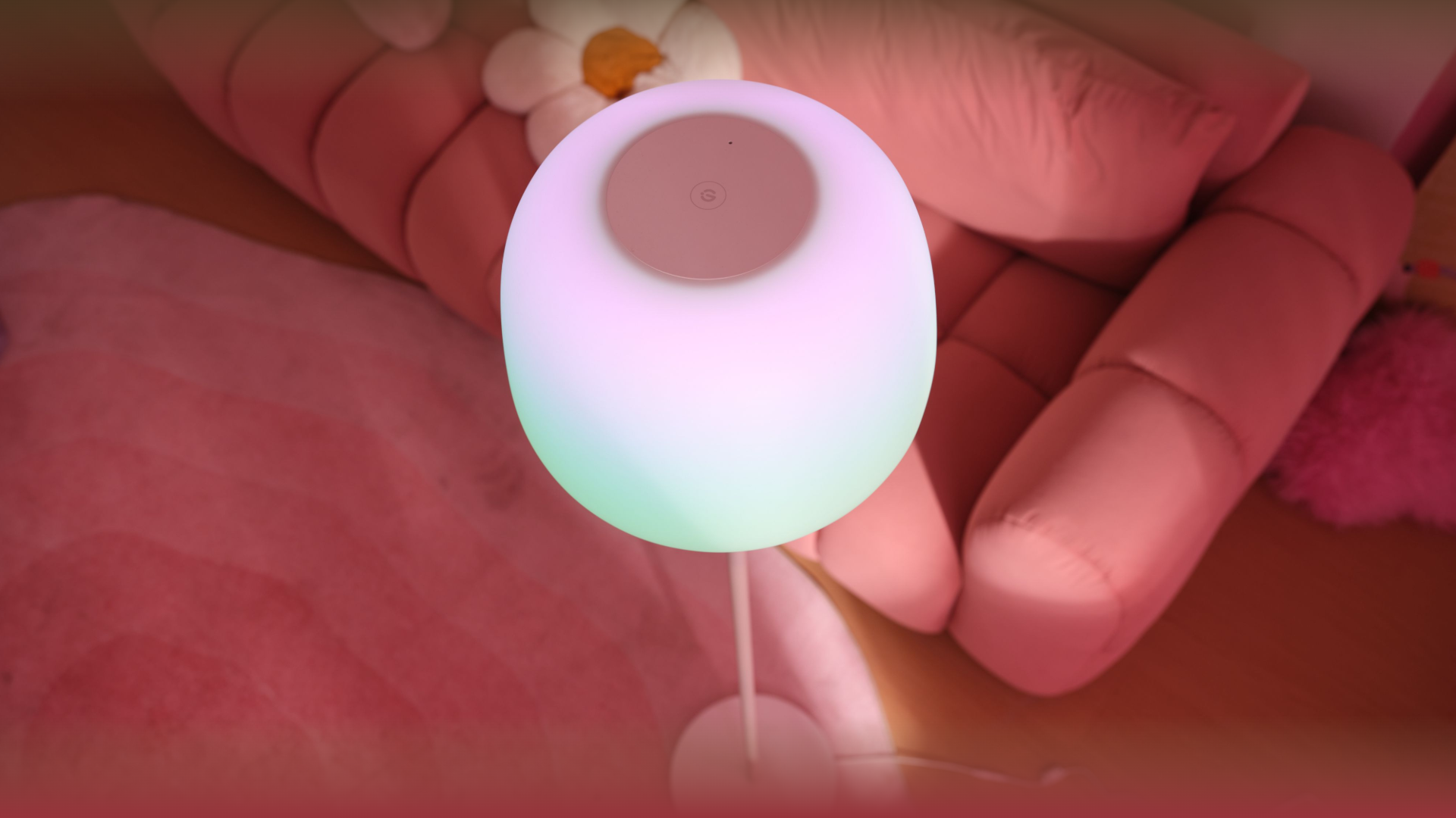 Eine runde, bunt leuchtende Stehlampe steht in einem Zimmer mit rosafarbenem Sofa und Teppich.