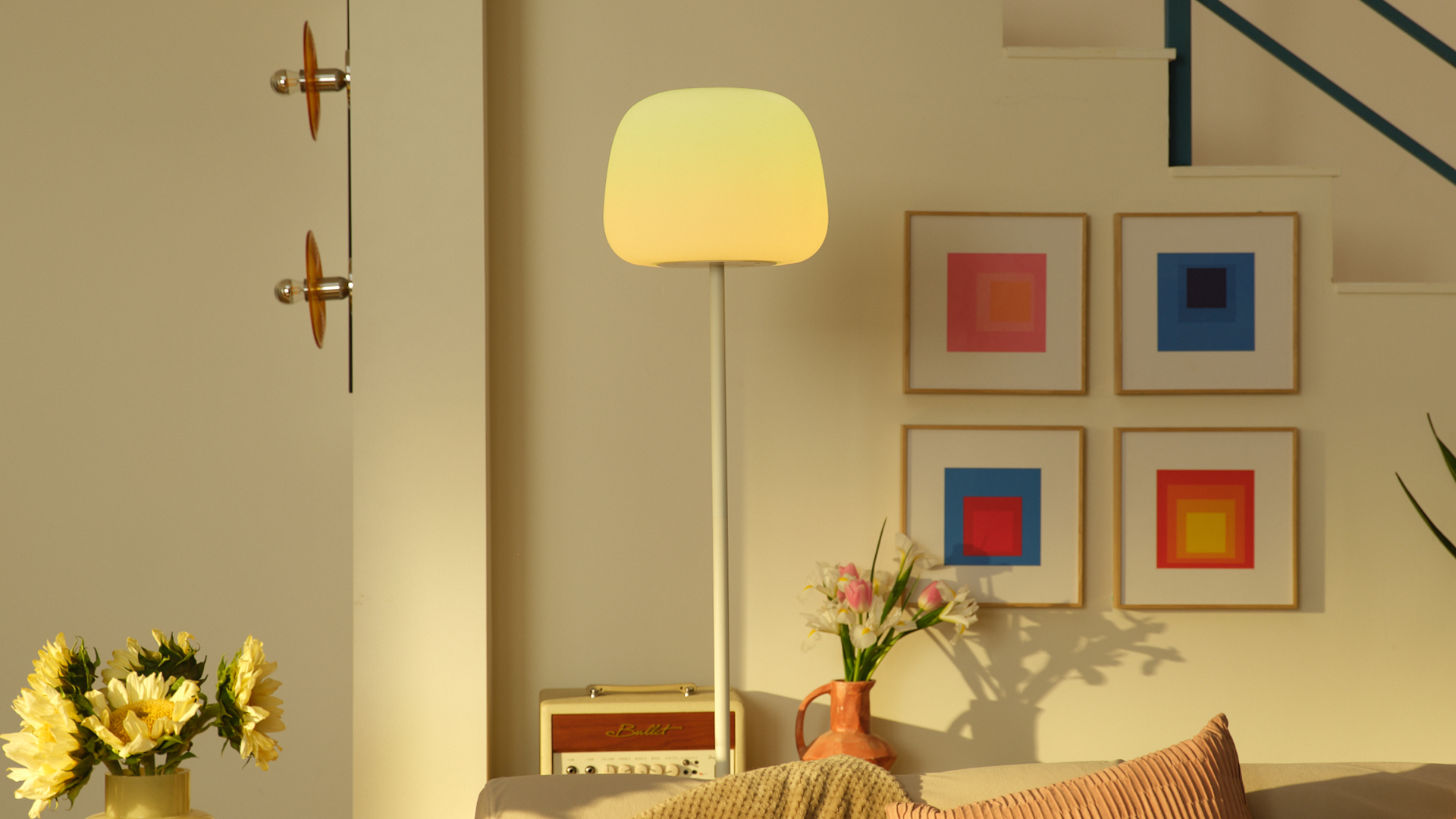 Beleuchtete Stehlampe in einem hellen Zimmer mit bunten Bildern, Blumen und einer Couch.
