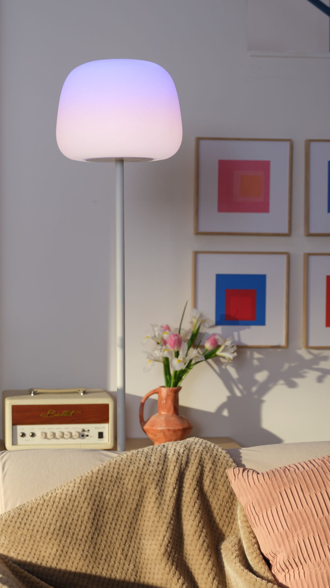 Eine Stehlampe mit blau-weißem Licht steht neben einem Sofa, im Hintergrund sind Bilder und Blumen zu sehen.