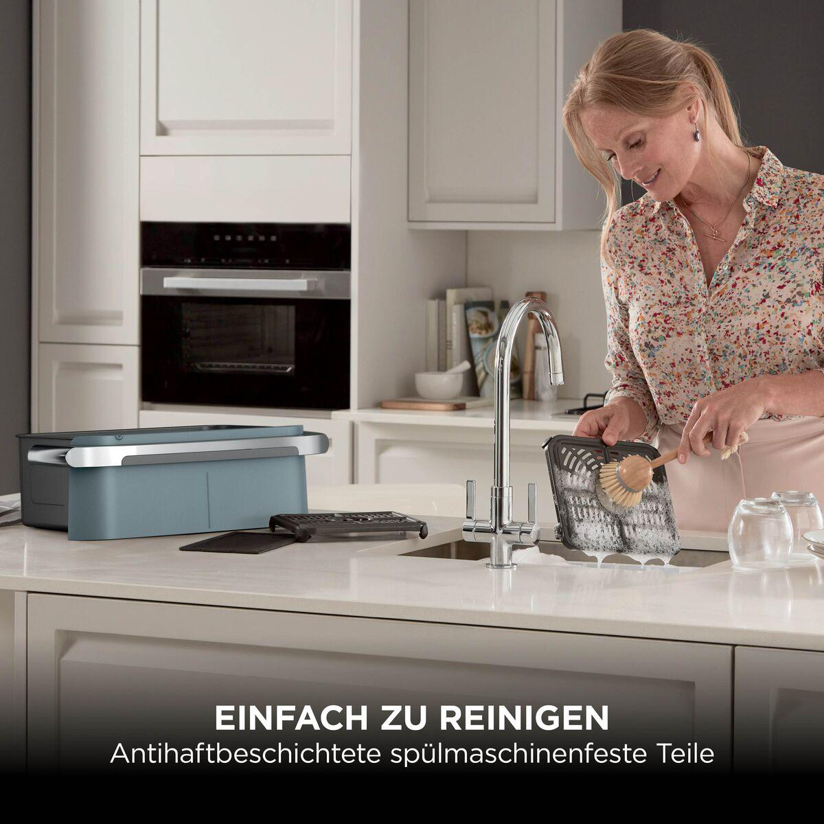 Eine Frau wäscht ein Küchenteil mit einer Bürste in einer Spüle. Im Hintergrund eine moderne weiße Küche.