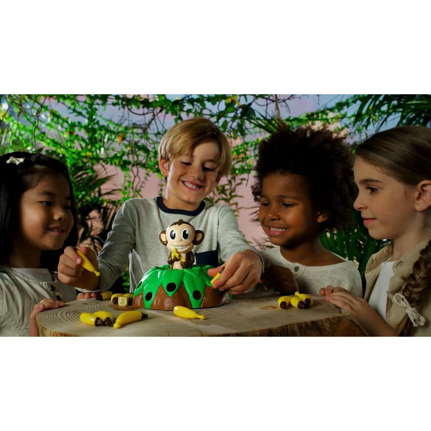 Quatre enfants souriants jouent à un jeu de singe sur une table en bois devant un feuillage vert.