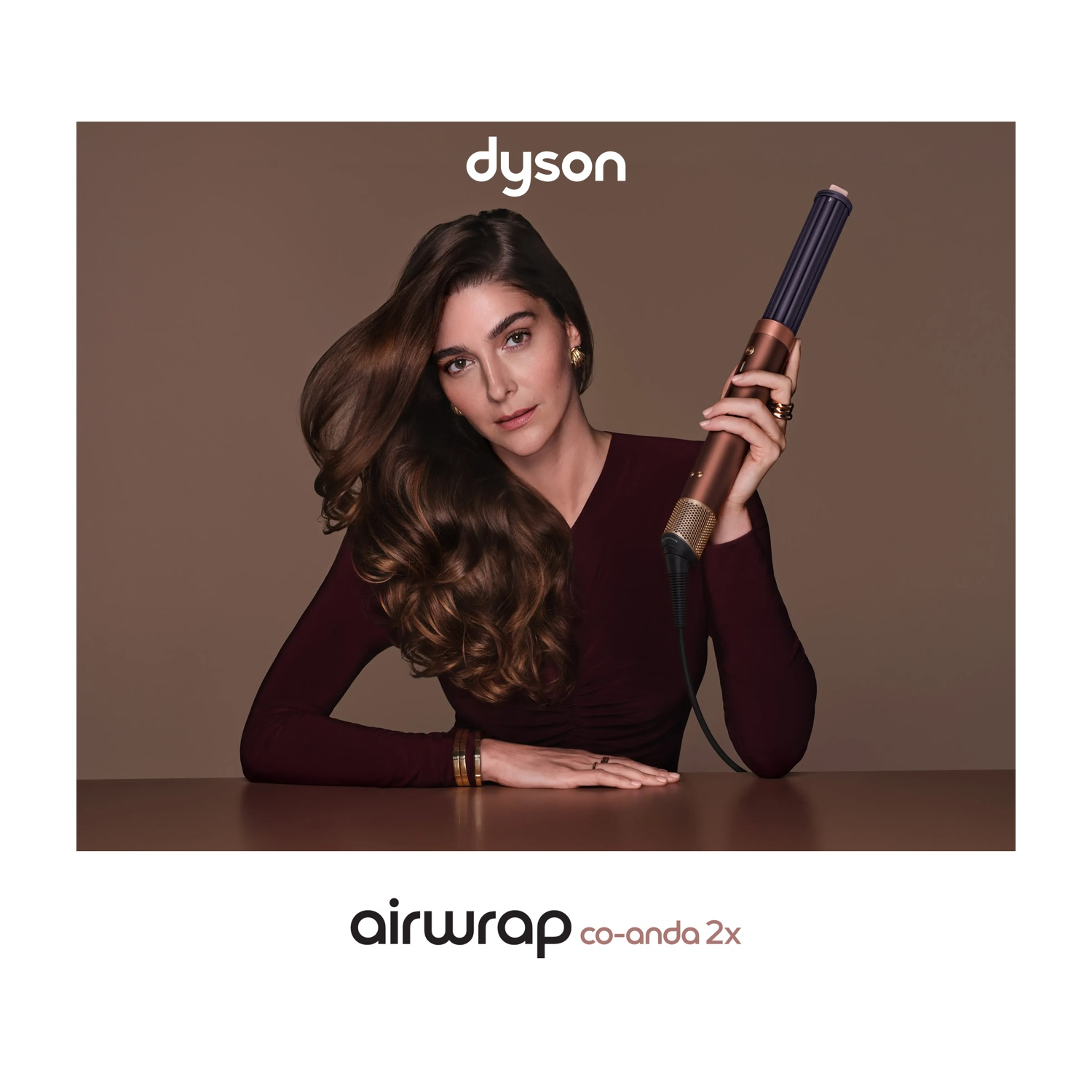 Una donna bruna con capelli ondulati tiene un Dyson Airwrap su sfondo marrone.